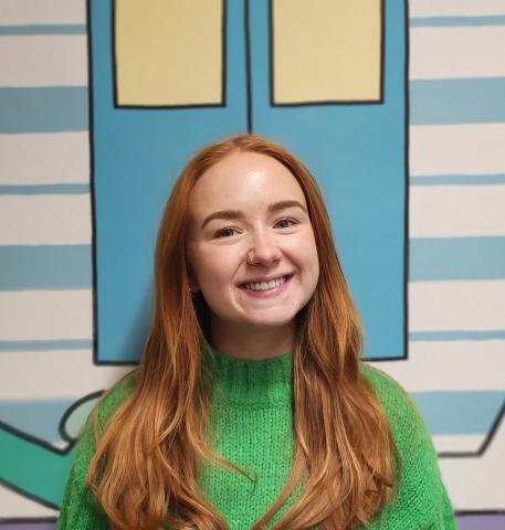 Amy standing in front of a pastel blue background, wearing a green jumper. 